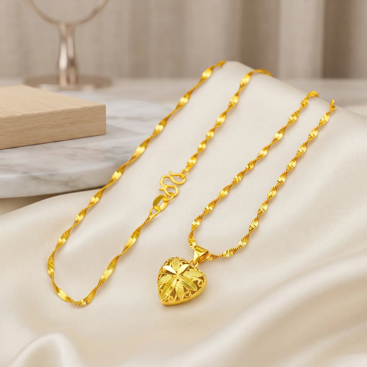 A gold necklace with a textured heart-shaped pendant, displayed on draped cream satin fabric with a marble block and soft neutral background.